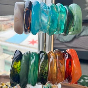 May include: A collection of sixteen resin bangle bracelets displayed on a vertical stand. The bracelets feature various translucent and opaque colors including white, black, turquoise, teal, green, amber, red, and orange with marbled patterns.