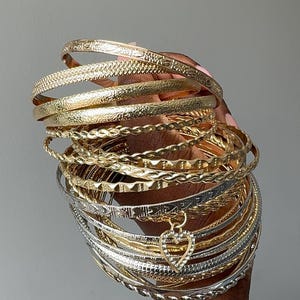 Op de afbeelding: Een verzameling gestapelde armbanden in goud- en zilverkleuren, met diverse texturen zoals gladde, gedraaide en bewerkte afwerkingen. Eén zilveren armband bevat een kleine, hartvormige bedel die is ingelegd met heldere, fonkelende steentjes.