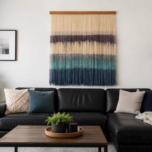 Puede incluir: Un tapiz de pared con largos hilos verticales de fibra suspendidos de una varilla de madera horizontal. El tejido presenta un patrón de color degradado que transiciona de crema en la parte superior a morado oscuro, turquesa y azul profundo.