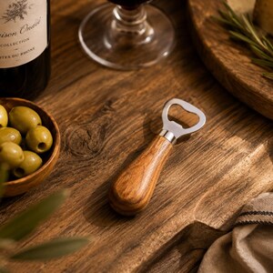 May include: A wooden-handled bottle opener with a silver head rests on a wooden surface. A bottle of wine, a bowl of olives, and a cheese board are also visible. The wine bottle label reads "Maison Ona&euml;l Collection C&ocirc;tes du Rh&ocirc;ne."