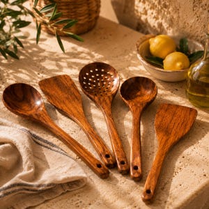 May include: A collection of five wooden kitchen utensils, including a spoon, spatula, slotted spoon, and ladle. The utensils are made of a rich, dark wood and are arranged on a light-colored surface. A bowl of lemons and olive oil are in the background.