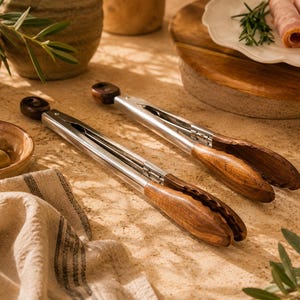 May include: Two stainless steel and wood serving tongs are displayed on a textured surface. The tongs have a polished metal body and dark wood handles. The image also includes olives, a wooden cutting board, and a linen cloth.