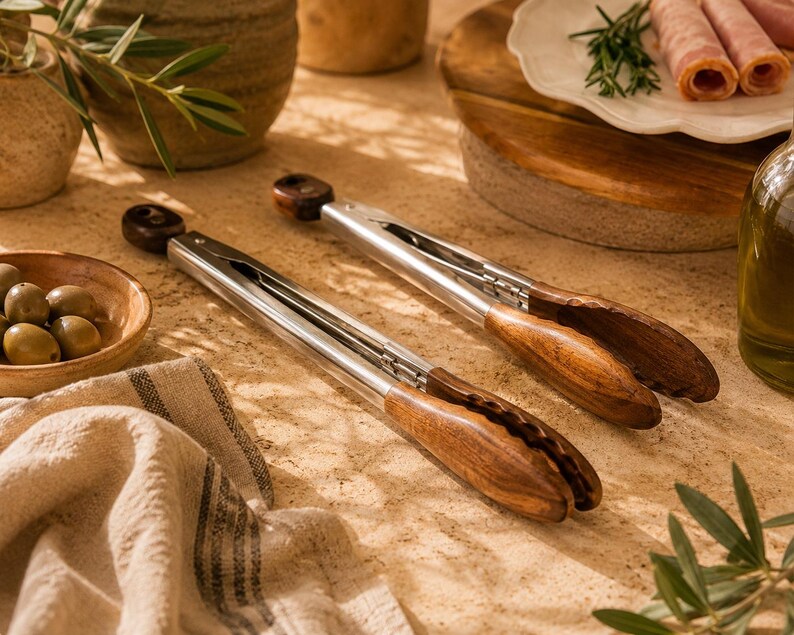 May include: Two stainless steel and wood serving tongs are displayed on a textured surface. The tongs have a polished metal body and dark wood handles. The image also includes olives, a wooden cutting board, and a linen cloth.