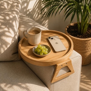 Könnte beinhalten: Ein runder, hellfarbiger Sofa-Arm-Tablett mit einer Kaffeetasse, einer kleinen Schale mit grünen Trauben und einem Smartphone. Das Tablett ist an der Armlehne eines beigen Sofas befestigt. Eine Topfpflanze befindet sich im Hintergrund.