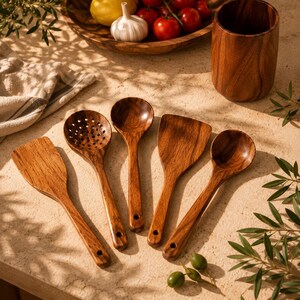 May include: A collection of wooden kitchen utensils, including a slotted spoon, spatula, and serving spoons. The utensils are made of a warm-toned wood and are arranged on a light-colored surface. A wooden cup and a bowl of fresh produce are in the background.