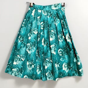 May include: A teal-colored skirt with a floral pattern of white roses. The skirt is pleated and hangs from a hanger. The fabric has a textured appearance, and the roses are scattered across the skirt.