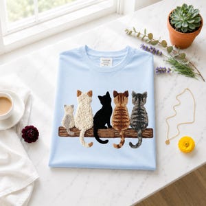 Può includere: Una t-shirt azzurro chiaro con un ricamo testurizzato raffigurante cinque gatti seduti in fila su un ramo di legno marrone. I gatti sono ritratti di schiena con manti di colore bianco, nero, tigrato marrone e tigrato grigio.