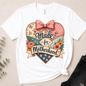 Peut inclure: Un t-shirt blanc à manches courtes orné d'un grand motif en forme de cœur avec un design patchwork dans les tons rose, bleu et crème. Le cœur est surmonté d'un nœud floral rose et porte l'inscription Made for Motherhood sur une bannière.