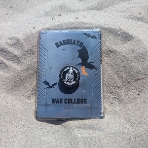 Puede incluir: Una funda rectangular para lector electrónico con el texto BASGIATH WAR COLLEGE, siluetas de dragones y espadas cruzadas. Un agarre circular para teléfono está fijado en el centro, mostrando una ilustración de esqueleto y el texto LEAVE ME ALONE I'M READING.