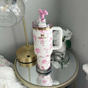 K&ouml;nnte beinhalten: Ein wei&szlig;er Stanley-Becher mit einem floralen Rosenmuster und einem rosa schleifenf&ouml;rmigen Deckel. Der Becher hat einen wei&szlig;en Griff und einen goldenen Rand. Das Stanley-Logo und "Loves Fancy" sind aufgedruckt.
