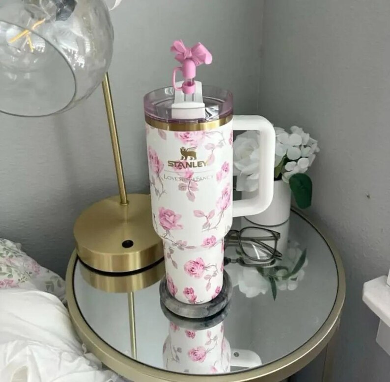 K&ouml;nnte beinhalten: Ein wei&szlig;er Stanley-Becher mit einem floralen Rosenmuster und einem rosa schleifenf&ouml;rmigen Deckel. Der Becher hat einen wei&szlig;en Griff und einen goldenen Rand. Das Stanley-Logo und "Loves Fancy" sind aufgedruckt.