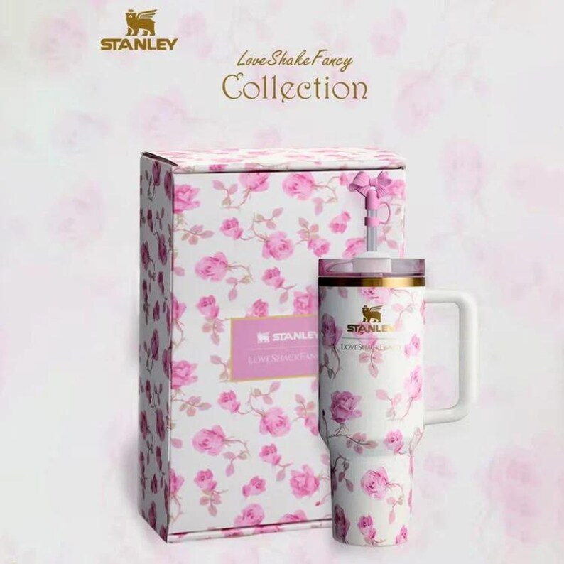 K&ouml;nnte beinhalten: Ein wei&szlig;er Stanley-Becher und eine passende Geschenkbox, beide mit rosa Rosenmustern verziert. Der Becher hat einen Griff und einen goldenen Rand. Der Text "LoveShakeFancy Collection" ist sichtbar.