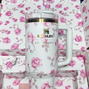 K&ouml;nnte beinhalten: Wei&szlig;er Stanley-Becher mit einem rosa Rosenbl&uuml;tenmuster, einem wei&szlig;en Griff und einem rosa schleifenf&ouml;rmigen Strohhalm-Aufsatz. Der Becher hat einen goldenen Rand und das Stanley-Logo. Die Verpackung weist ebenfalls das gleiche florale Design auf.