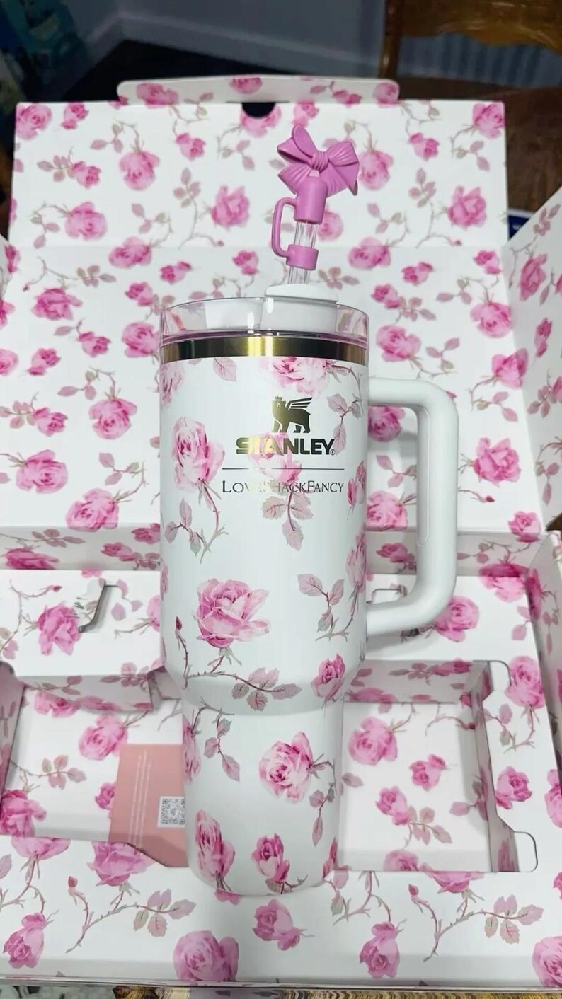 K&ouml;nnte beinhalten: Wei&szlig;er Stanley-Becher mit einem rosa Rosenbl&uuml;tenmuster, einem wei&szlig;en Griff und einem rosa schleifenf&ouml;rmigen Strohhalm-Aufsatz. Der Becher hat einen goldenen Rand und das Stanley-Logo. Die Verpackung weist ebenfalls das gleiche florale Design auf.