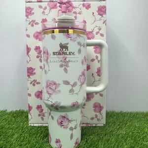 K&ouml;nnte beinhalten: Ein wei&szlig;er Stanley-Reisebecher mit einem rosa Rosenmuster und einem gro&szlig;en Griff. Der Becher hat einen rosa Deckel und einen goldenen Rand. Der Text "Stanley" und "LoveShackFancy" ist auf der Vorderseite aufgedruckt. Eine passende Box befindet sich hinter dem Becher.