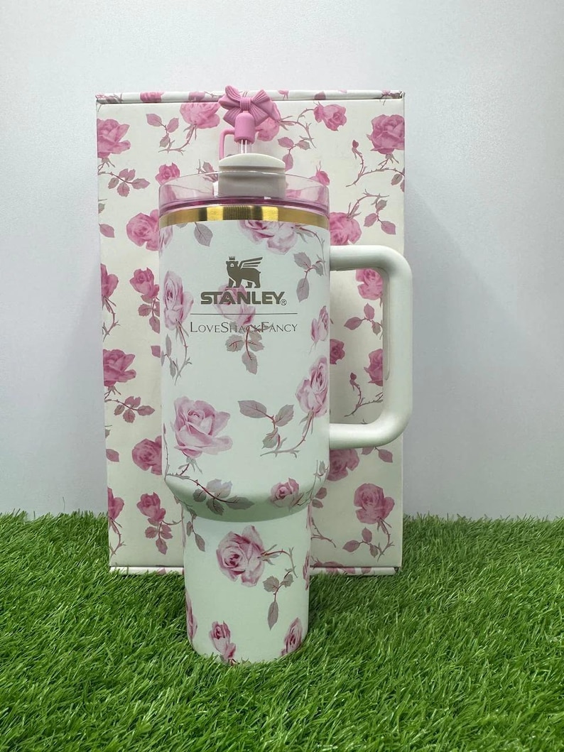 K&ouml;nnte beinhalten: Ein wei&szlig;er Stanley-Reisebecher mit einem rosa Rosenmuster und einem gro&szlig;en Griff. Der Becher hat einen rosa Deckel und einen goldenen Rand. Der Text "Stanley" und "LoveShackFancy" ist auf der Vorderseite aufgedruckt. Eine passende Box befindet sich hinter dem Becher.
