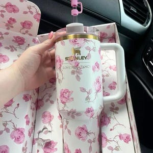 K&ouml;nnte beinhalten: Wei&szlig;er Stanley-Becher mit rosa Rosenmuster, goldfarbenem Rand und wei&szlig;em Griff. Der Becher befindet sich in einer passenden floralen Box und hat einen rosa schleifenf&ouml;rmigen Strohhalmaufsatz. Der Text "Stanley Loves Fancy" ist sichtbar.