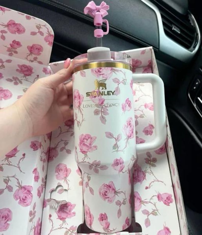 K&ouml;nnte beinhalten: Wei&szlig;er Stanley-Becher mit rosa Rosenmuster, goldfarbenem Rand und wei&szlig;em Griff. Der Becher befindet sich in einer passenden floralen Box und hat einen rosa schleifenf&ouml;rmigen Strohhalmaufsatz. Der Text "Stanley Loves Fancy" ist sichtbar.