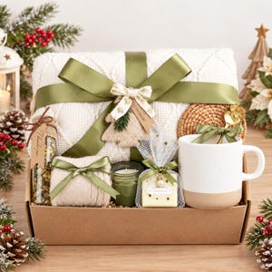 Könnte beinhalten: Eine Geschenkbox mit einer weißen Strickdecke, die mit einem grünen Band gebunden ist, einer weißen Keramiktasse mit goldenem Löffel, einer kleinen grünen Kerze, beigen Socken, einem Glas mit Trockenblumen und einer in weißem Netz verpackten Seife.