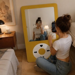 May include: A large, rectangular, mustard yellow floor mirror designed to resemble a vintage digital music player. The mirror features a white circular control interface at the bottom with black icons for playback, including a heart and arrows.