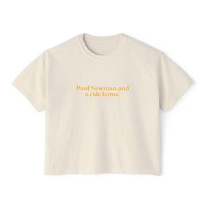 May include: Cream-colored t-shirt with the text "Paul Newman and a ride home" in orange. The shirt has short sleeves and a crew neck. The text is centered on the front of the shirt.