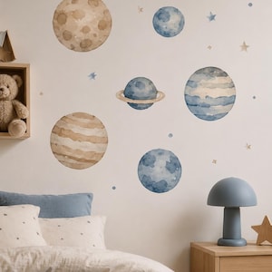 Könnte beinhalten: Ein Set aus Aquarell-Wandaufklebern mit verschiedenen Planeten in Blau- und Beigetönen, darunter ein Planet mit Ringen sowie kleine verstreute Sterne und Punkte, die auf einer schlichten Wand über einem Bett angeordnet sind.