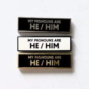 Puede incluir: Tres pines de esmalte con fondos blanco y negro. Cada pin tiene el texto "MY PRONOUNS ARE HE/HIM" en letras plateadas, blancas y doradas.