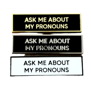 May include: Three black and white enamel pins with gold, silver, and white metal frames. Each pin reads "Ask me about my pronouns".