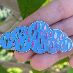 May include: A blue enamel pin shaped like a cloud with colourful raindrops. The raindrops are in various shades of pink, purple, blue, and white.