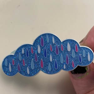 Peut inclure: Une épingle en forme de nuage bleu pailleté avec des gouttes de pluie blanches, bleues et roses.
