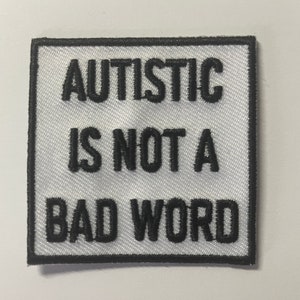 May include: A square, white patch with black embroidered text that reads "AUTISTIC IS NOT A BAD WORD." The patch has a black border and is likely designed to be sewn onto clothing or accessories.
