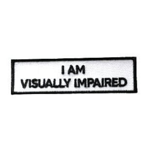 Op de afbeelding: Een rechthoekige witte patch met zwarte geborduurde tekst die luidt: "I AM VISUALLY IMPAIRED." De patch heeft een zwarte rand en staat tegen een witte achtergrond.