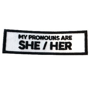 Puede incluir: Parche rectangular blanco con borde negro y texto que dice "My pronouns are she/her".
