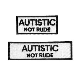 Könnte beinhalten: Zwei rechteckige Aufnäher in Schwarz und Weiß. Die Aufnäher tragen den Text "AUTISTISCH NICHT UNHÖFLICH" in schwarzen Blockbuchstaben. Der Text ist innerhalb eines weißen Rechtecks zentriert, das von einem schwarzen Rechteck umrandet ist. Die Aufnäher befinden sich auf weißem Hintergrund.