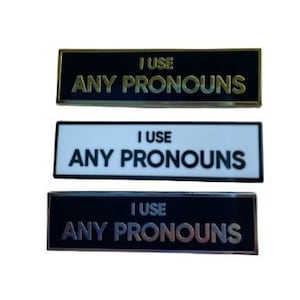 May include: Three black rectangular name tags with gold trim. Each tag reads "I use any pronouns". The top tag has a black background, the middle tag has a white background, and the bottom tag has a light brown background.