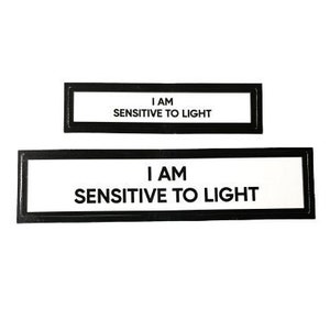 May include: Two black framed signs with white backgrounds. The top sign reads "I AM SENSITIVE TO LIGHT". The bottom sign reads "I AM SENSITIVE TO LIGHT".