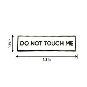 May include: A rectangular enamel pin with a white background and a black border. The text DO NOT TOUCH ME is printed in black capital letters. The pin measures 1.5 inches in width and 0.39 inches in height as indicated by the measurement lines.