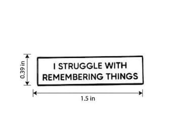 May include: A rectangular white enamel pin with a black border and black text that reads I STRUGGLE WITH REMEMBERING THINGS. The pin measures 1.5 inches in width and 0.39 inches in height as indicated by the visible measurement lines and labels.