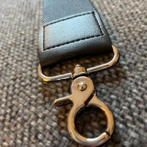 Puede incluir: Primer plano de una correa negra con un cierre de metal plateado. La correa está hecha de un material texturizado y está unida al cierre con una pieza de cuero cosida. El cierre tiene un gancho giratorio.