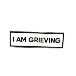 May include: A white enamel pin with a black border. The pin reads "I AM GRIEVING" in black text.