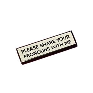 May include: A black and white enamel pin with the text "PLEASE SHARE YOUR PRONOUNS WITH ME" on a white background.