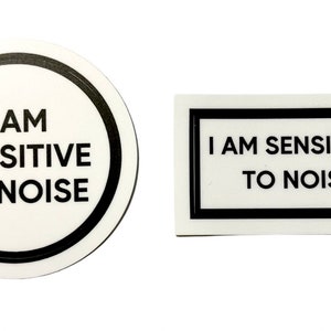 May include: Two white stickers with black text and borders. One sticker is round and says "I AM SENSITIVE TO NOISE". The other sticker is rectangular and says "I AM SENSITIVE TO NOISE".