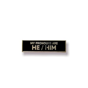 May include: Black enamel pin with silver trim. The pin reads "My pronouns are He/Him".