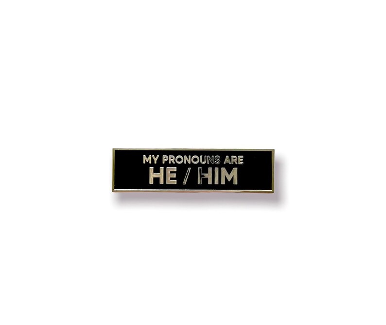 May include: Black enamel pin with silver trim. The pin reads "My pronouns are He/Him".