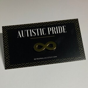 May include: A black card with gold trim and the text "AUTISTIC PRIDE" in white. A gold infinity symbol is centered on the card. The text "RETROPHILIA.ETSY.COM" is at the bottom of the card.