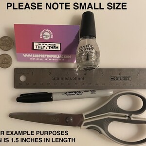 Puede incluir: Primer plano de un peque&ntilde;o pin con el texto "My pronouns are they/them" sobre un fondo rosa y blanco. El pin est&aacute; al lado de una regla, un marcador negro, unas tijeras y una botella de esmalte de u&ntilde;as transparente. El texto "Please note small size" est&aacute; en la parte superior de la imagen. El texto "For example purposes pin is 1.5 inches in length" est&aacute; en la parte inferior de la imagen.