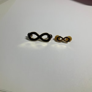 May include: Two infinity symbols, one black and one gold, are displayed on a white surface. The black infinity symbol is larger than the gold one.