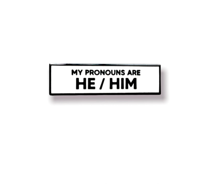 May include: Black and white enamel pin with the text "My pronouns are He/Him".
