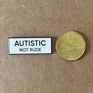 May include: A white rectangular button with black text that reads "AUTISTIC NOT RUDE". The button is next to a Canadian one dollar coin.