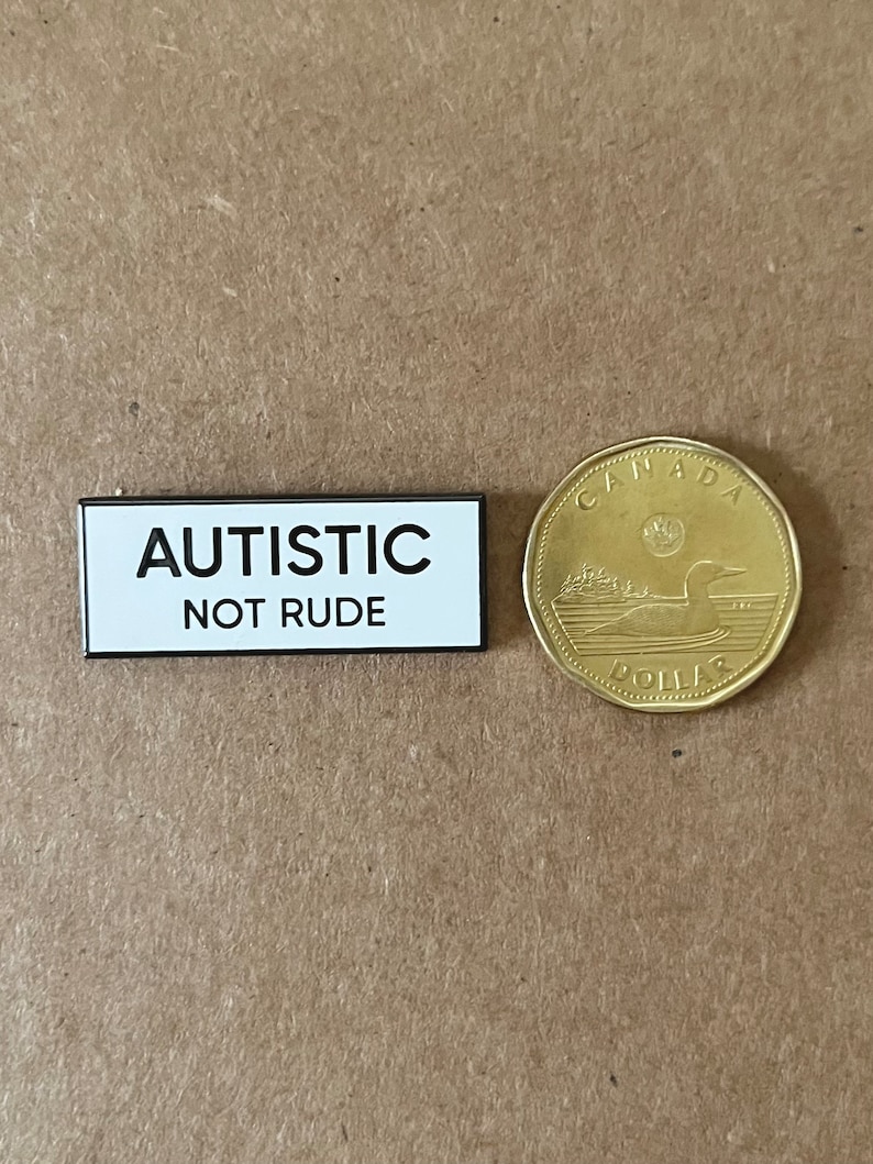 May include: A white rectangular button with black text that reads "AUTISTIC NOT RUDE". The button is next to a Canadian one dollar coin.
